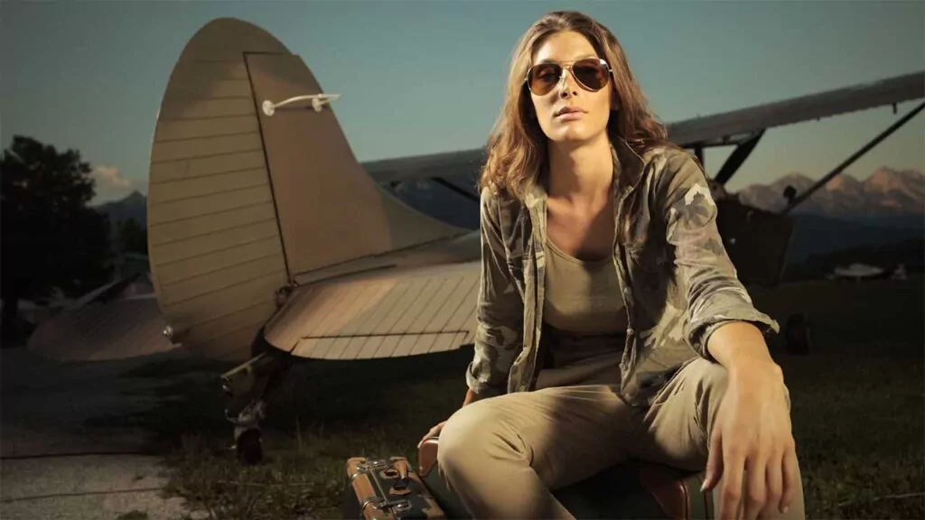 Woman in aviator sunglasses sitting by a vintage aircraft, holding a suitcase