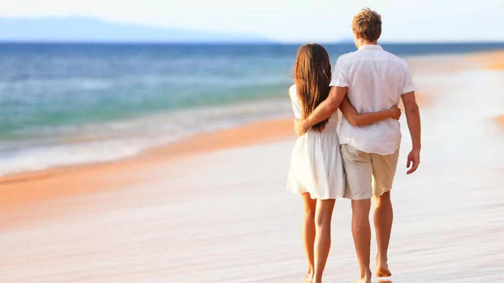 Couple walking arm in arm on a sandy beach.